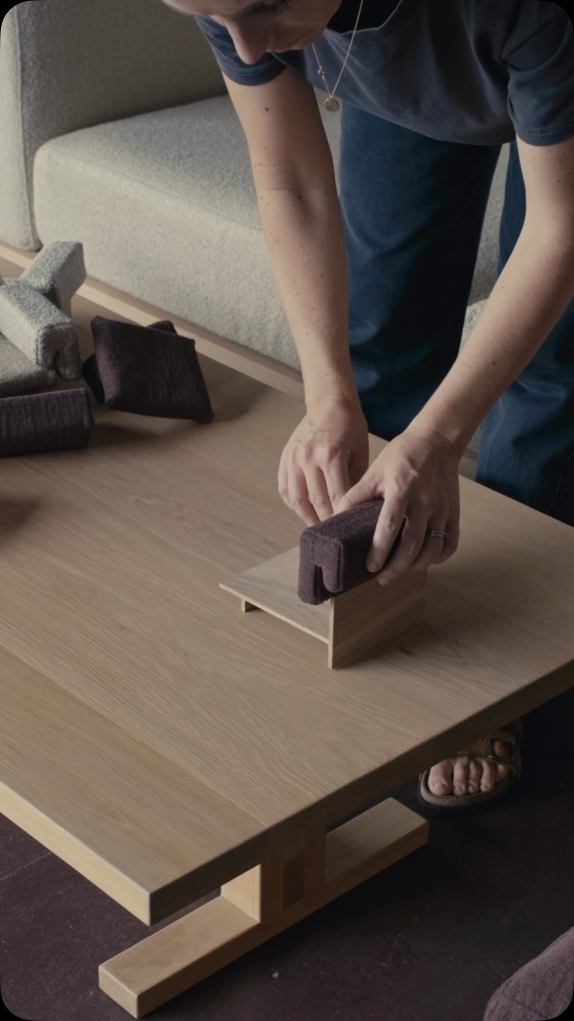 The image shows a woman's hands manipulating small furniture pieces over a wooden table. Several mini cushions and blocks are arranged on the table, alongside a larger sofa piece in the background. The scene suggests a playful setup or demonstration of modular furniture.