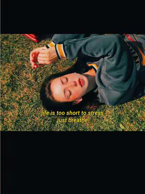 A young woman lies on grass, looking up with her eyes closed. She wears a gray hoodie with yellow accents. Text overlayed in a yellow font reads, "life is too short to stress, just breathe."