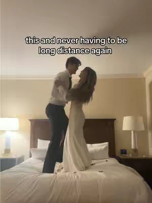 A couple is embracing, jumping on a bed. The man is lifting the woman into the air as they embrace. The setting is a hotel room or bedroom, with a bed, nightstands, and lamps visible. The walls are a neutral color, and the lighting appears warm.