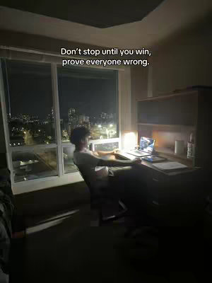 A dimly lit room with a large window showcasing a night cityscape. A person is seated at a desk, partially visible, with a laptop and papers, illuminated by a desk lamp. Text overlay is visible in a white font, reading 'Don't stop until you win, prove everyone wrong.'