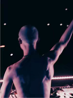 A shirtless man from the back, holding his arm up with an excited hand. The shot is from a live event with dark setting lights in the background, focused on the energetic atmosphere.