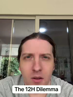 A selfie-style video cover image featuring a person with light skin, dark hair, and light-colored eyes looking directly at the camera with a neutral expression. The person is wearing a gray shirt. The background is blurred, with a window and a glimpse of the outside visible. A white rectangular text box at the bottom of the image reads 'The 12H Dilemma' in sans-serif font.