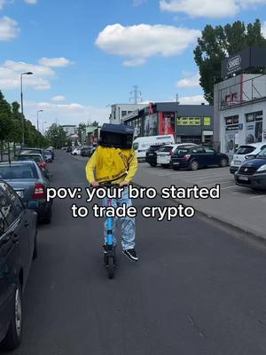 A person wearing a yellow jacket and a box on the head rides an electric scooter down a road. The person is riding on the left side of the road, and there are parked cars on each side. The sky is blue with some clouds, and the background includes buildings and trees.