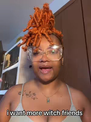 A medium shot of a woman with orange locs, wearing large clear-framed glasses and a light green tank top. She is looking at the camera and speaking. The background is a neutral-toned space, possibly a room in a house. Text overlayed at the bottom says 'i want more witch friends' in white.