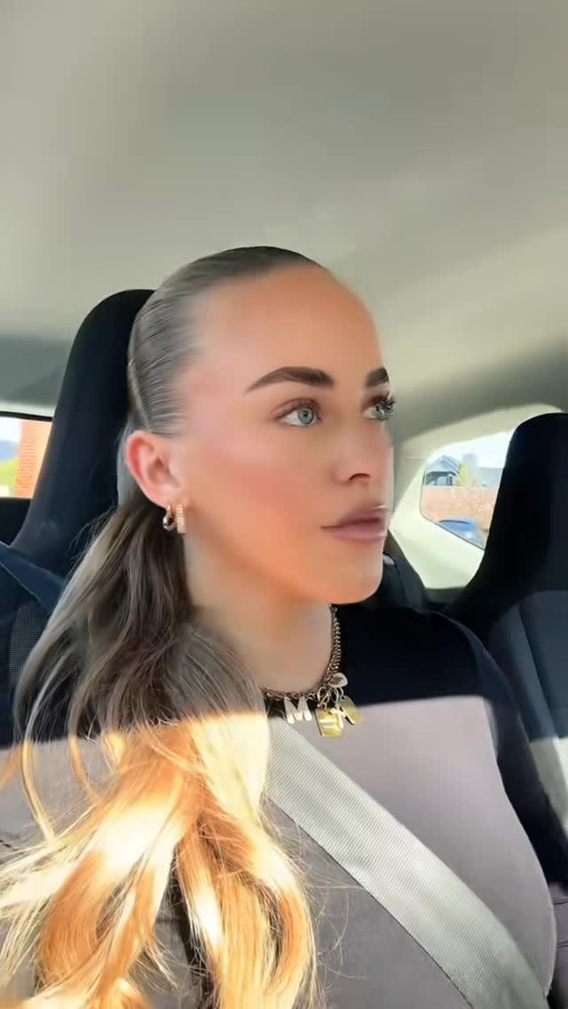 A woman is sitting in a car, looking out the window. She has blonde hair pulled back in a ponytail and is wearing a black top and a necklace. The lighting is bright, and the overall composition is a selfie-style shot.