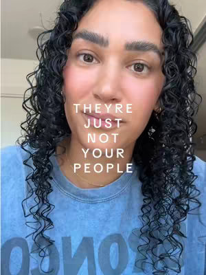 A selfie-style shot featuring a woman with curly dark hair and brown eyes, centered against a soft, neutral backdrop. Text overlayed on the image reads 'THEY'RE JUST NOT YOUR PEOPLE' in a clean, sans-serif font, stacked vertically. She is wearing a blue t-shirt. The overall lighting is natural and soft, emphasizing the woman's features and the message of the overlay.