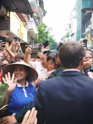 The image captures a crowd of people, many holding up phones to take photos. In the foreground, a woman in a blue shirt and sun hat waves at the camera with a big smile. Behind her, a man, presumably the subject, is partially visible, facing away from the camera. The setting appears to be a street in a city, with buildings lining the sides.