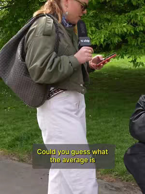A woman wearing sunglasses, a green jacket, and white pants is holding a microphone and a phone while on a sidewalk, with a green background.