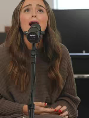 A woman with long brown hair is singing into a microphone. She is wearing a brown sweater. The background is blurred, though a dark object can be seen behind her.