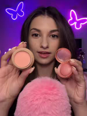 A woman is centered in the frame, holding two blush compacts. She has long brown hair and is looking directly at the camera. The background is a purple hue with two illuminated butterfly outlines. In the foreground, a pink fluffy texture sits below the woman’s hands.