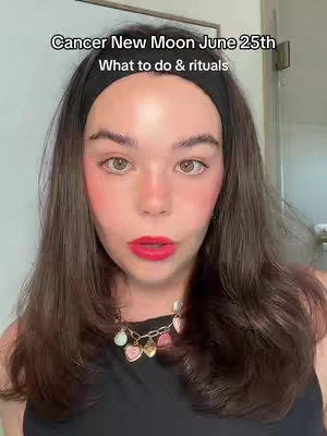 A woman with fair skin and long, dark brown hair is the focus of the image. She has prominent blush on her cheeks and bright red lipstick. She wears a black headband and a black top. A necklace with colorful heart-shaped pendants is visible. The background appears to be a room, with a blurred image of a door or mirror.