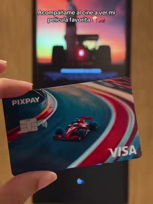 A hand holding a Pixpay Visa card, with the card's design featuring a red F1 race car on a track. In the background, a phone screen shows an F1 car silhouette and text that reads 'Acompáñame al cine a ver mi película favorita'. The overall composition has a warm lighting.