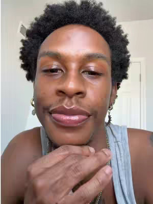 A close-up shot of a person sticking their tongue out. The person appears to be a Black male with short, dark, curly hair and is wearing a gray tank top. They have multiple earrings, and a gold chain is visible. The background is a white wall with a door frame.