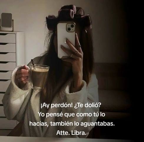 A selfie-style image featuring a person holding a smartphone and a glass of coffee. The person is wearing a bathrobe and has curlers in their hair, suggesting a relaxed, at-home setting. The background includes a white dresser and a mirror. Text is overlaid on the image, written in Spanish, with an implied sarcastic tone, and the text is focused on a Libra zodiac sign.