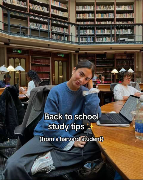 A young woman in a blue sweater sits in a library, leaning on her left hand and with her legs crossed. She is wearing grey pants, a white shirt underneath, and silver shoes. Behind her are bookshelves filled with books, and other students can be seen in the background studying. The image is well-lit and has a warm color palette.