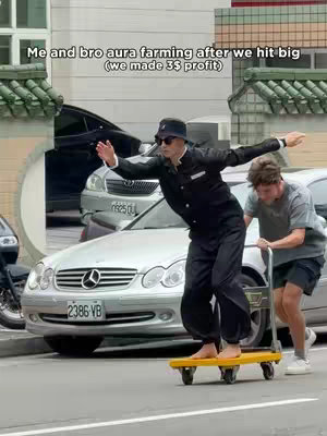 An image of two men in a city street. One man, dressed formally in a black suit and bucket hat is balanced on a small yellow cart, arms outstretched, with a look of mock seriousness. A second man pushes the cart from behind. In the background are parked cars on a street, with buildings and green foliage visible. Text is overlaid on the image.