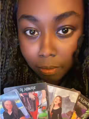 A close-up image of a person, likely a woman, with dark skin, large brown eyes, and braided hair. She is holding a variety of tarot cards, the central focus of the image. The cards are held in front of her face, partially obscuring it. The overall lighting is soft, with a warm tone, possibly from indoor lighting.