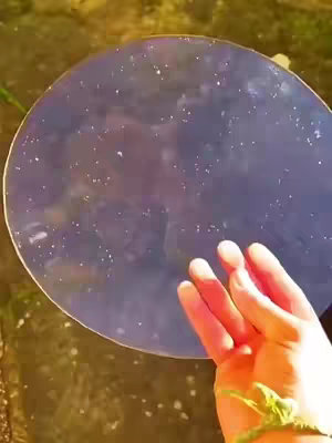 A hand with a slightly reddish complexion reaches towards a large, circular, reflective surface, likely water. The surface reflects a dark, purplish hue interspersed with small, white speckles, creating a starry night effect. The background features a soft, out-of-focus area with muted yellow and green tones, suggesting an outdoor setting. The composition offers a calming and potentially intriguing visual experience.