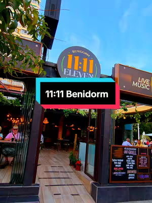 An outdoor shot featuring a restaurant entrance in Benidorm. A black bar with the words "11:11 Benidorm" in white text on a red background is overlayed at the bottom. The image shows a restaurant called "11:11 Eleven" with a sign above the entrance. People are dining outdoors. The image has a slightly warm color palette with a blue sky.