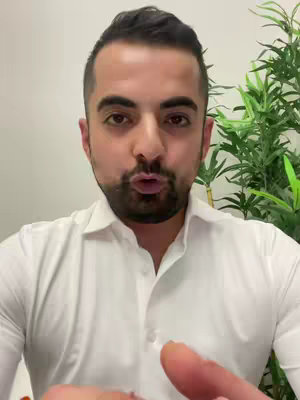 A man with dark hair and a beard is speaking directly to the camera. He is wearing a white button-down shirt, and a background of greenery - possibly a plant - is visible to his right. His mouth is open as if he is speaking. The image is well lit, with the man’s face being the primary focus. The composition is a medium shot, including both his face and upper chest.