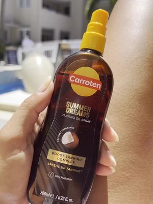 A person holds a bottle of Carroten tanning oil against a sunny background, with the bottle taking up most of the frame. The bottle is brown and has a yellow cap. The person's skin and part of a background pool are visible on the right side. The Carroten logo and product details are clearly displayed on the bottle.
