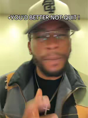 A man wearing glasses, a baseball cap, a black shirt, and a jacket is looking at the camera. The image has warm lighting and is slightly blurred.