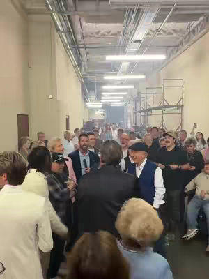 A behind-the-scenes shot of what appears to be a backstage area or hallway. A large group of people is visible, and in the foreground, several individuals are congregated around a man holding a microphone, presumably warming up his voice. The setting is brightly lit with overhead lights, and a long series of conduit and metal structures run along the ceiling, giving a sense of depth. The overall composition is candid with a focus on multiple people in an active setting.