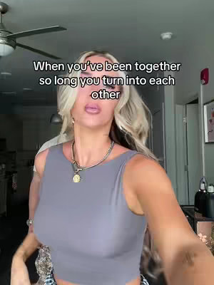 A woman stands in a well-lit room, with the camera close to her. She wears a gray top and a necklace with a circular pendant. Her blonde hair frames her face, and the background is slightly out of focus, suggesting a casual, indoor setting.