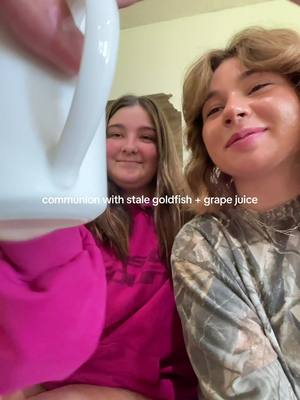 Two young women, one holding a white pitcher, pose for a selfie. The woman holding the pitcher is on the left, looking at the camera with a smile. The other woman is on the right, smiling slightly. The background is a lightly colored room, with text overlayed on the image stating "communion with stale goldfish + grape juice."