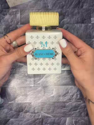 A close-up shot of a white perfume bottle held by two hands with manicured white nails. The bottle is rectangular with a light yellow wooden cap. The bottle has a light blue label with 'BLANC CRÉME' written on it, and a stylized letter 'M'. The background is a textured gray surface.