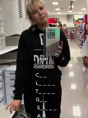 A person is holding a book in a retail store, with text overlaid on the image that makes a word puzzle. The person wears a black hoodie and stands amid shopping aisles, including shelves, offering context and suggesting a lifestyle or shopping theme.