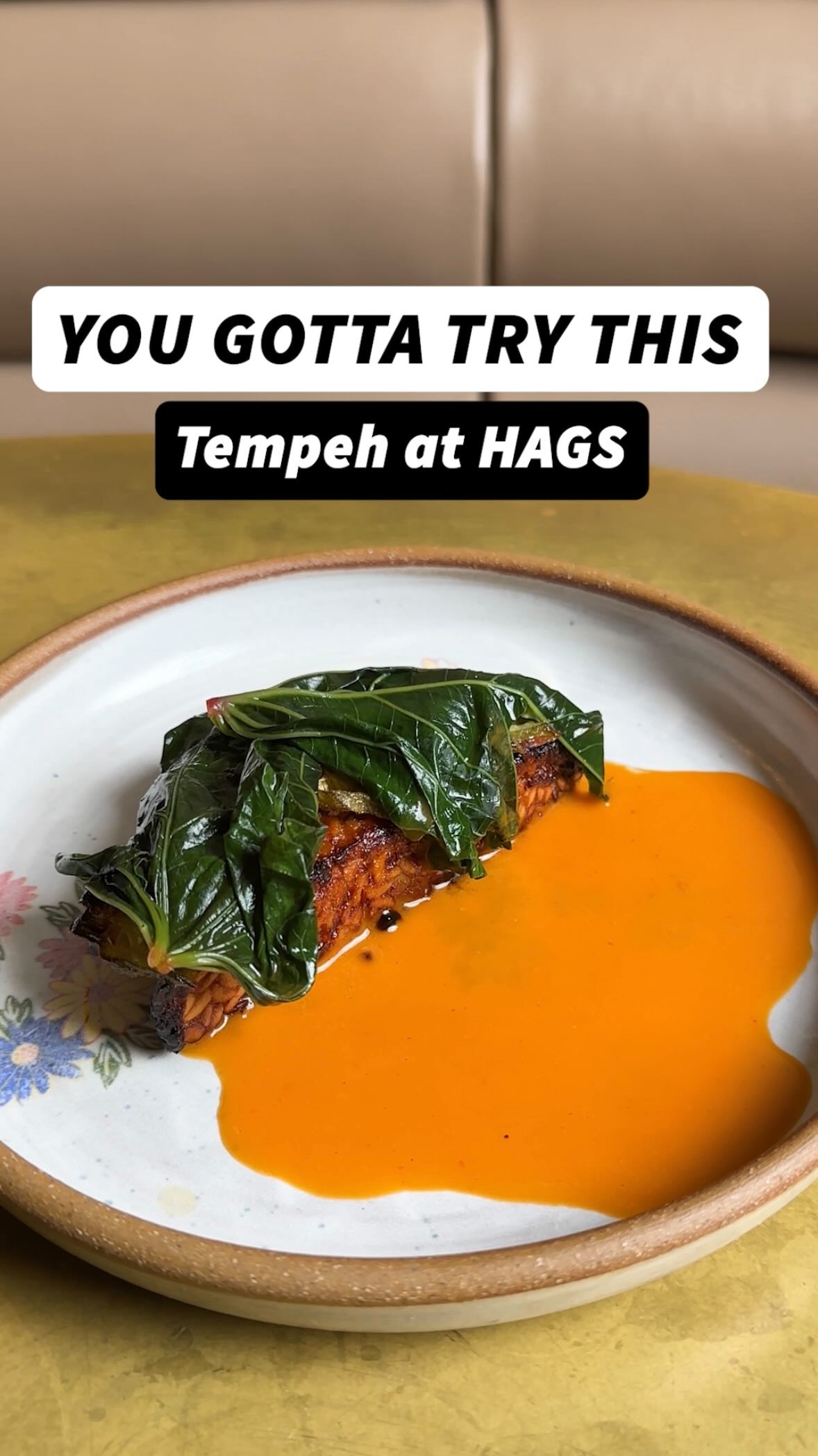 A close-up shot of a plated dish, featuring a square-shaped tempeh covered in large green leaves, resting on a vibrant orange sauce. The plate has a floral patterned rim. The background is blurred, suggesting an interior setting.