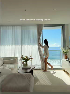 A bright, airy interior shot features a room with large windows opening to a view of the ocean. A woman stands near the window, holding the curtain. The room is decorated in neutral colors, with white furniture, a floral arrangement on the coffee table, and sunlight streaming in through the window. The overall impression is one of elegance, tranquility, and a dream lifestyle.