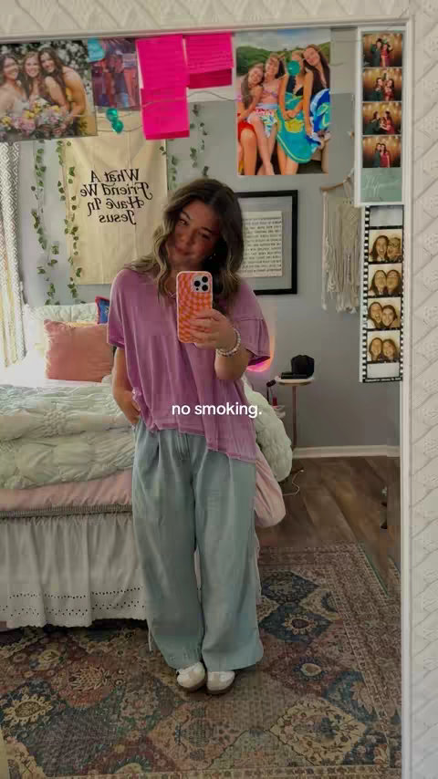 A woman takes a mirror selfie in a bedroom with decorative elements. She wears a purple shirt and loose, light blue jeans. A phone with an orange patterned case obscures her face. The mirror reflects a room with a bed, posters, and personal items.