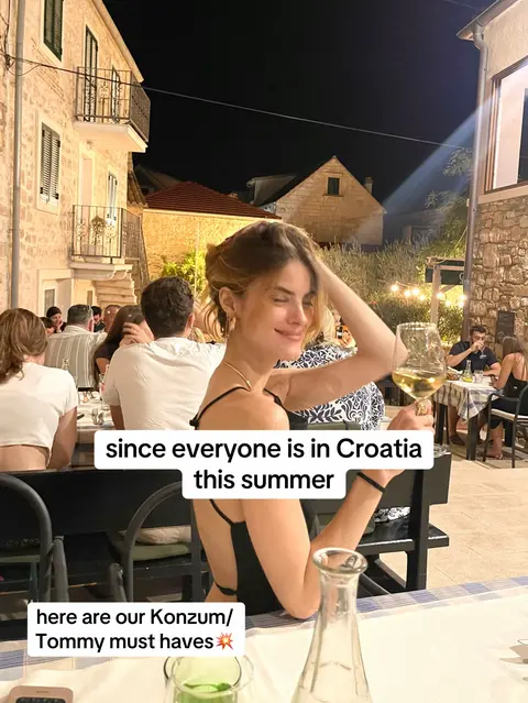 A woman with blonde hair in a black top is seated at a table outdoors. She has a glass of white wine in her hand, smiling with her eyes closed. Behind her is a stone building and other people seated at tables.