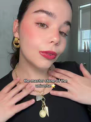 A close-up selfie of a woman with brown eyes and dark hair. She has red blush on her cheeks and glossy red lipstick. She is wearing gold hoop earrings and a black shirt. Gold chain necklace with charm pendant is visible, with the text overlayed on the image.
