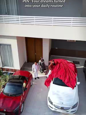 An overhead shot of a luxurious house, driveway occupied by a red sports car and a white car. People are present in the setting. Text appears at the top of the image.