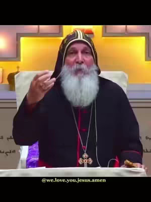 A medium shot features an older man, likely a religious figure, speaking. He wears a black robe with a red lining at the collar and a head covering with an ornate cap. A large, white beard covers most of his lower face. He is gesturing with his hand, and a decorative gold necklace with a cross hangs around his neck. The background includes a yellow panel behind him and the environment appears to be illuminated in a warm tone. At the bottom of the frame is text containing a username.