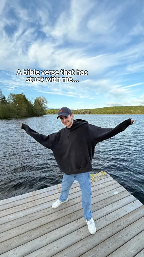 A man stands on a wooden dock with his arms outstretched, seemingly hugging the scene of a lake and trees. Text overlayed includes the phrase 'A bible verse that has stuck with me...'. The background features a calm lake under a blue sky with scattered clouds.