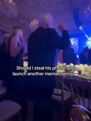 A dimly lit scene at a lavish event, possibly a wedding or gala. A person in a dark suit is seen from behind, with their head and shoulders visible. Other figures, including a woman in a black dress, are situated in the background, along with tables adorned with white floral arrangements. The overall color scheme is dark with pops of light, indicative of a festive atmosphere.