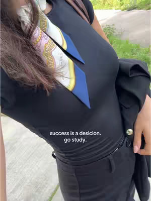 An over-the-shoulder shot of a person dressed in a black top and pants. The top is form-fitting, and a blazer is draped over the person's arm. A scarf is worn around the neck with accents of gold and royal blue. The person looks to be walking on a stone path.