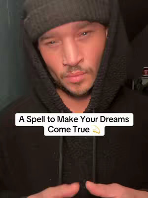 A man wearing a dark gray beanie and a black hooded sweatshirt looks down, seemingly introspective. A white rectangular text overlay near the bottom reads, 'A Spell to Make Your Dreams Come True.' The background is a blurry, dark interior.
