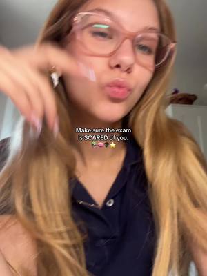 A young woman with long blonde hair and pink glasses, wearing a dark blue collared shirt, puckers her lips at the camera. A text overlay states, 'Make sure the exam is SCARED of you,' decorated with a small graphic of a candy, a flower, a graduation cap, and a star.