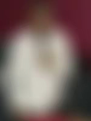 The image is a blurred portrait. Details are obscured by the blur, but a figure with a light-colored top is visible against a gradient background, transitioning from dark red or maroon at the top to black at the sides.