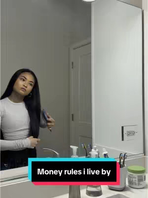 A woman is standing in a bathroom, looking in the mirror while holding a hairbrush and brushing her hair. A text overlay in a rectangular box at the bottom reads 'Money rules i live by'.