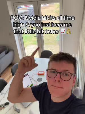 A selfie-style shot of a young man indoors, pointing upwards with his right hand. He has short, dark hair, glasses, and is wearing a dark blue t-shirt. The background includes a white table, a window, and a glimpse of outdoor greenery. Text overlayed on the image reads 'POV: Nvidia hit its all time high & you just became that little bit richer' with a rising graph and a money bag icon. There is also a small orange device on the table.