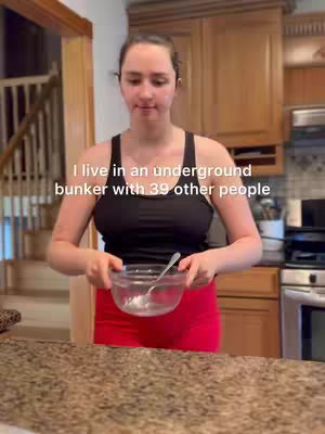 A woman stands in a kitchen setting, holding a glass bowl. The background includes wooden cabinets and a staircase. Text overlayed on the image reads, 'I live in an underground bunker with 39 other people'.