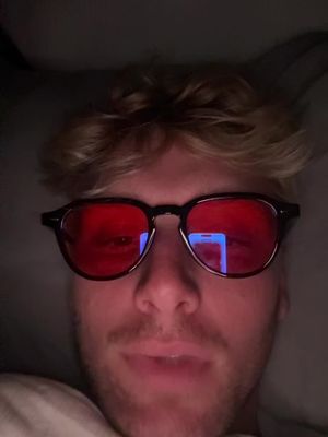 A selfie-style shot featuring a young man wearing dark-framed glasses with red lenses. He has light-colored hair and a slight smile. The lighting is dim, suggesting a low-light environment, with the red lenses reflecting bright light sources, possibly a smartphone screen. The background is out of focus and dark.