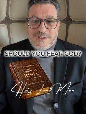 The cover image is a selfie-style shot of a man against a blurred background. The man, who appears to be the creator, is wearing glasses, a dark suit jacket, and a light-colored turtleneck. He is holding a closed book, which is the central focus, titled "THE ORIGINAL BIBLE", in front of him that is slightly angled towards the viewer, placed slightly below his face. Text overlayed on the image reads, 'SHOULD YOU FEAR GOD?' The background is out of focus, and beneath the book is the handwritten signature, “Holy Land Man”. The color palette is muted, with the man's skin tone and book adding contrast to the darker clothing.