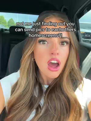 A selfie taken from inside a car, featuring a young woman with long, light brown hair and a surprised expression. Her mouth is open, and her eyes are wide. The background is blurred, suggesting movement or being out, with a glimpse of green trees visible. The upper text overlay reads, 'not me just finding out you can send pics to eachothers homescreens?!'.